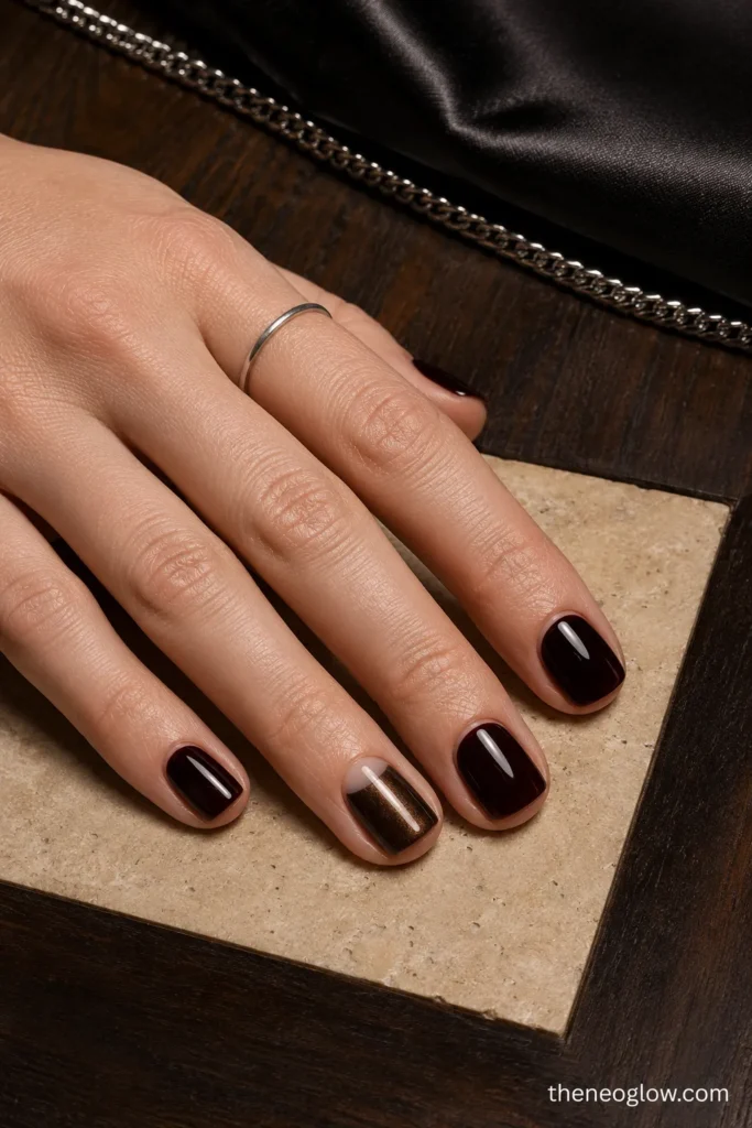 Black-cherry short square nails with one espresso and bronze-chrome half-moon accent beside a black satin clutch.