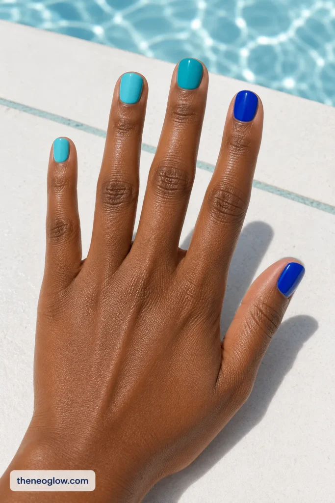 Short squoval nails alternating glossy cobalt, aqua, and turquoise on deep brown skin beside pool tile.