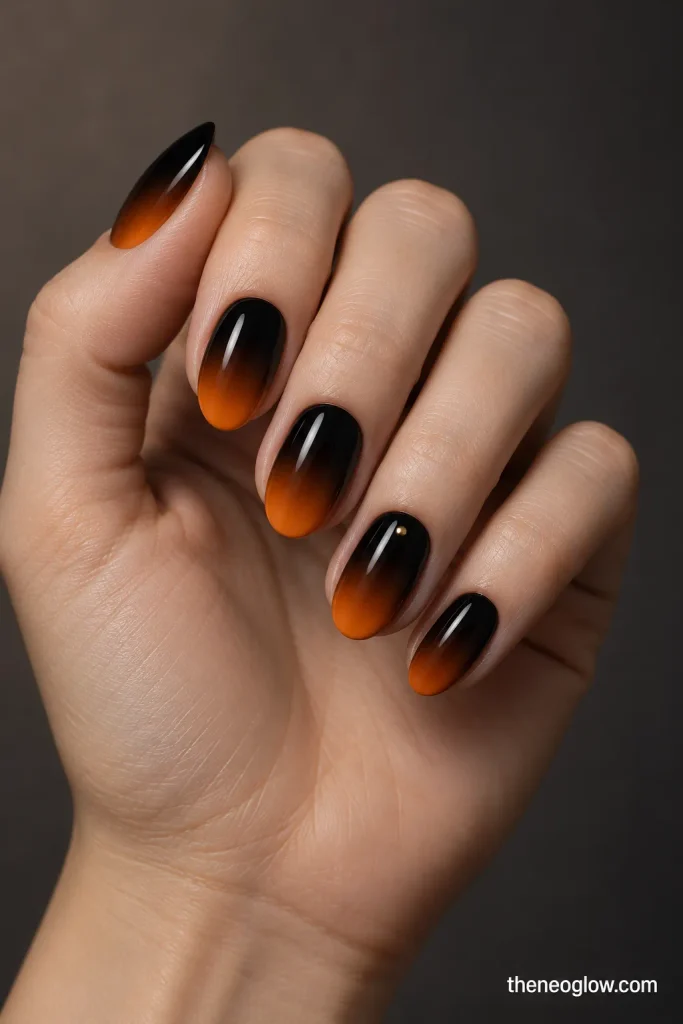 Medium oval nails fading from glossy black into burnt orange with one tiny gold pinpoint near a cuticle. black and orange Halloween nails