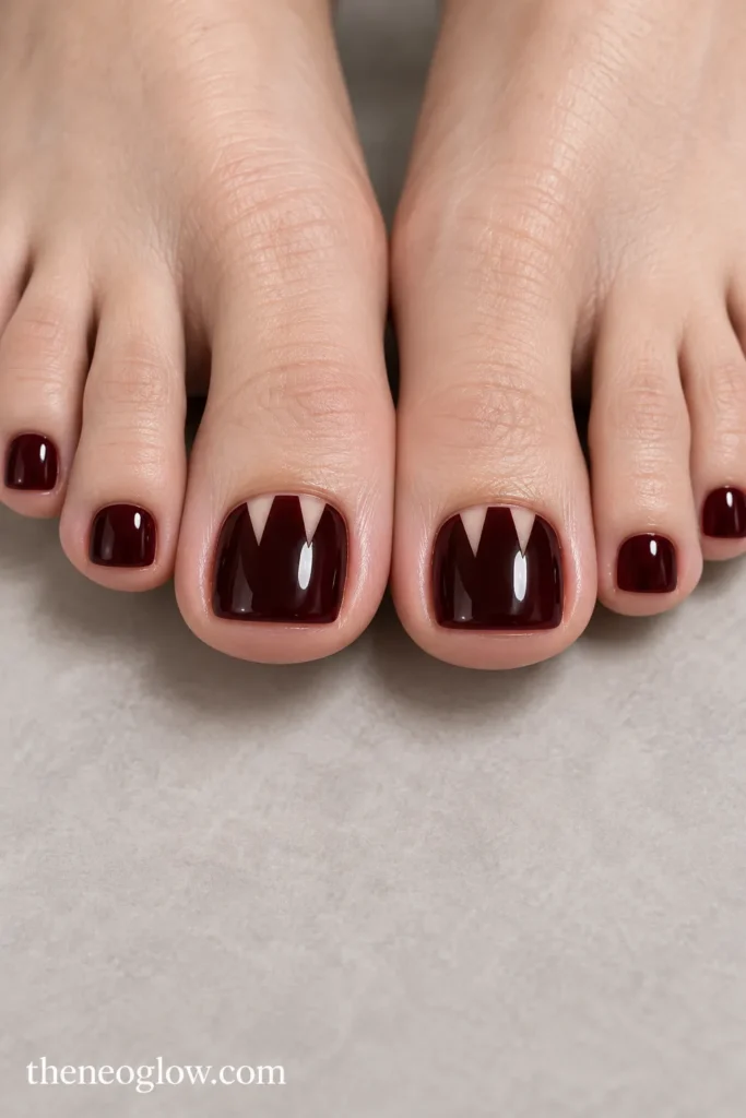 Deep black-cherry pedicure with two pointed negative-space fang cutouts on the big toenail
