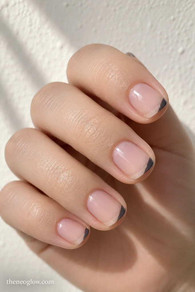 Very short sheer squoval nails with tiny charcoal polish confined to one outer tip corner.