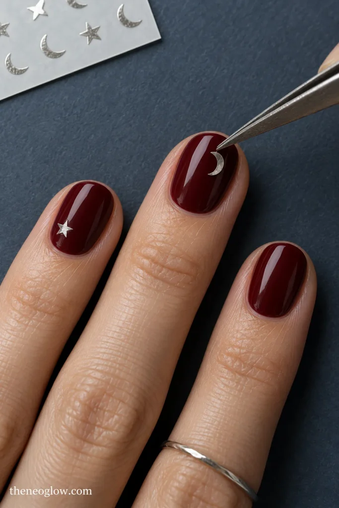 Tweezers place a small silver crescent sticker on a burgundy short oval nail beside a tiny star accent.