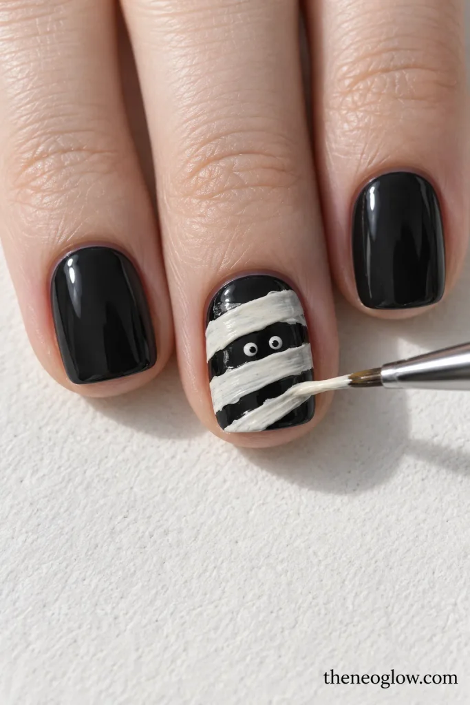 A fine brush paints off-white mummy bands over a charcoal short square accent nail with two tiny dark eyes.