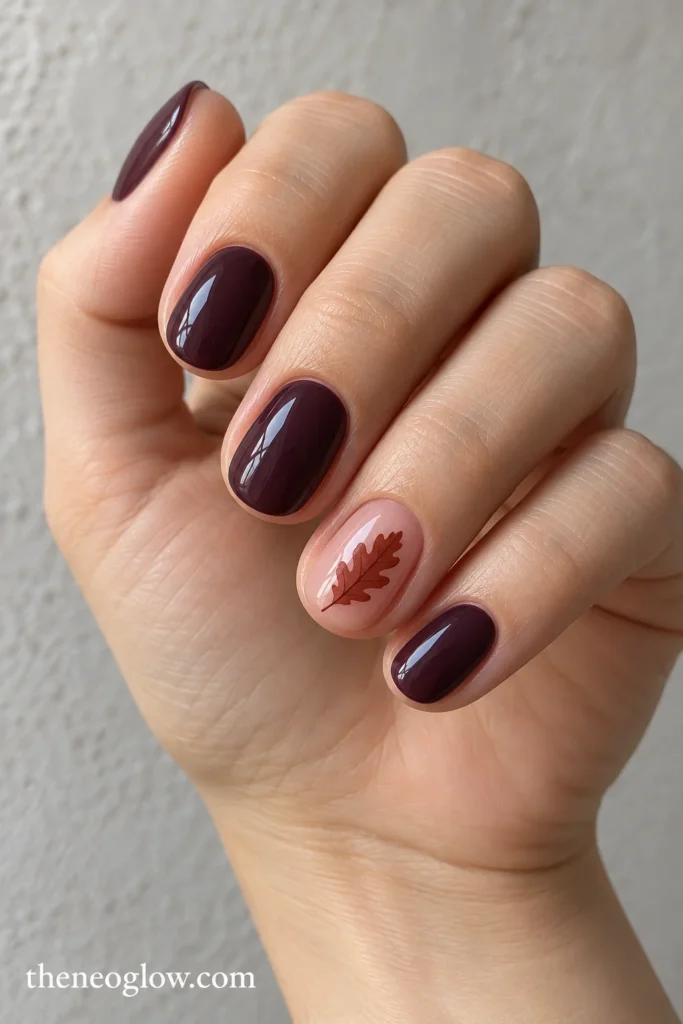 Glossy aubergine oval nails with one rosy transparent accent and a dark-rust oak leaf.