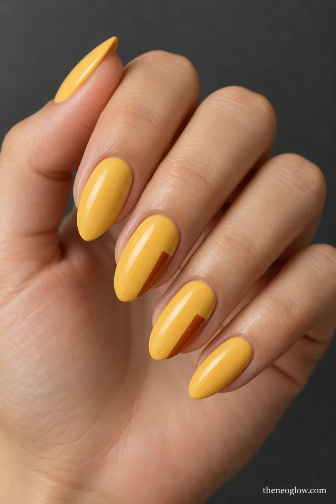 Muted yellow almond nails with slim burnt-sienna side panels on two fingers.