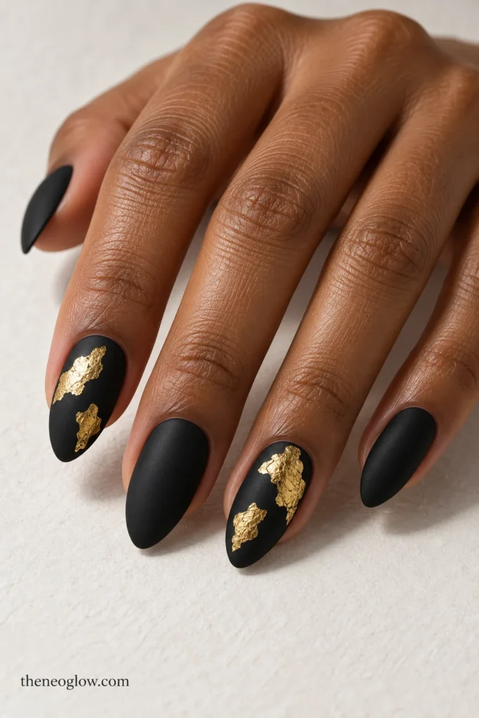 Medium matte black almond nails with irregular flat antique-gold foil pieces on the index and ring fingers.