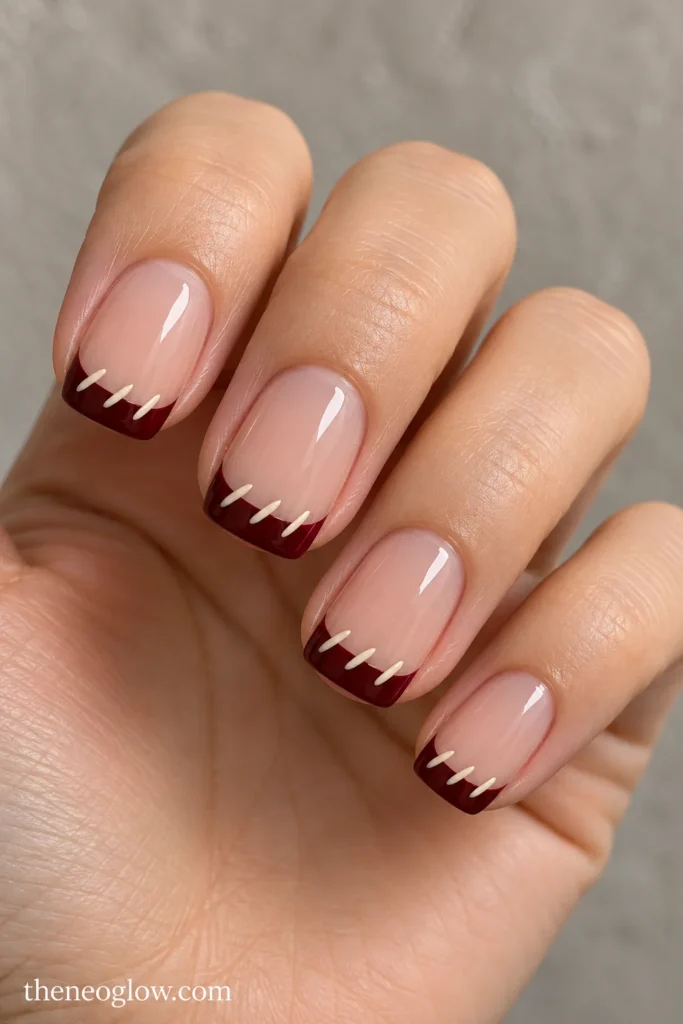 Medium squoval nails with wine-colored French tips and three tiny cream diagonal stitches along each inner edge.