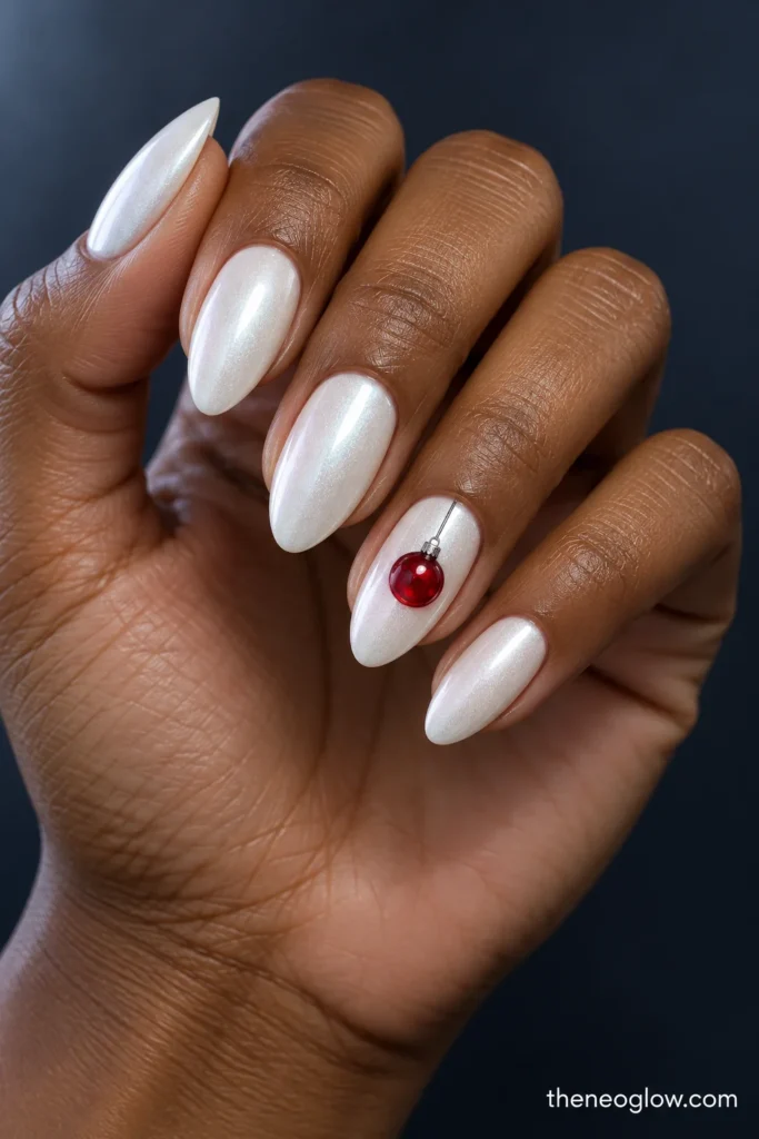 Pearly-white almond nails featuring a small ruby-red Christmas bauble hanging from a thin silver thread on the ring finger.